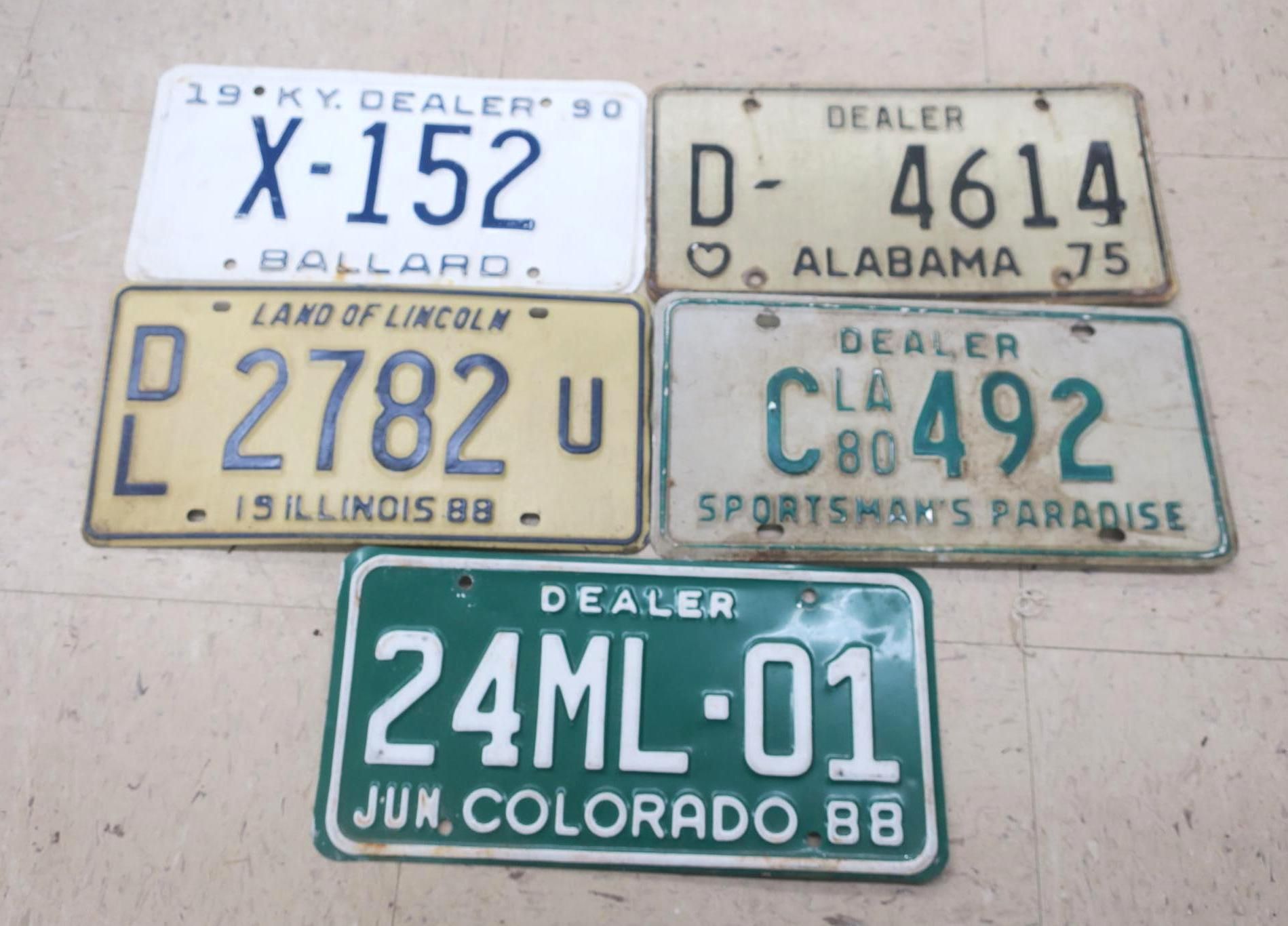 Five vintage dealer license plates arranged in a grid, in blue, tan, yellow, teal, and green.