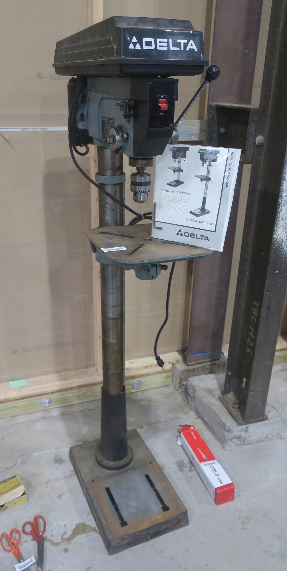 Delta drill press with a white paper hanging from the table in a workshop corner, next to a wall and floor.
