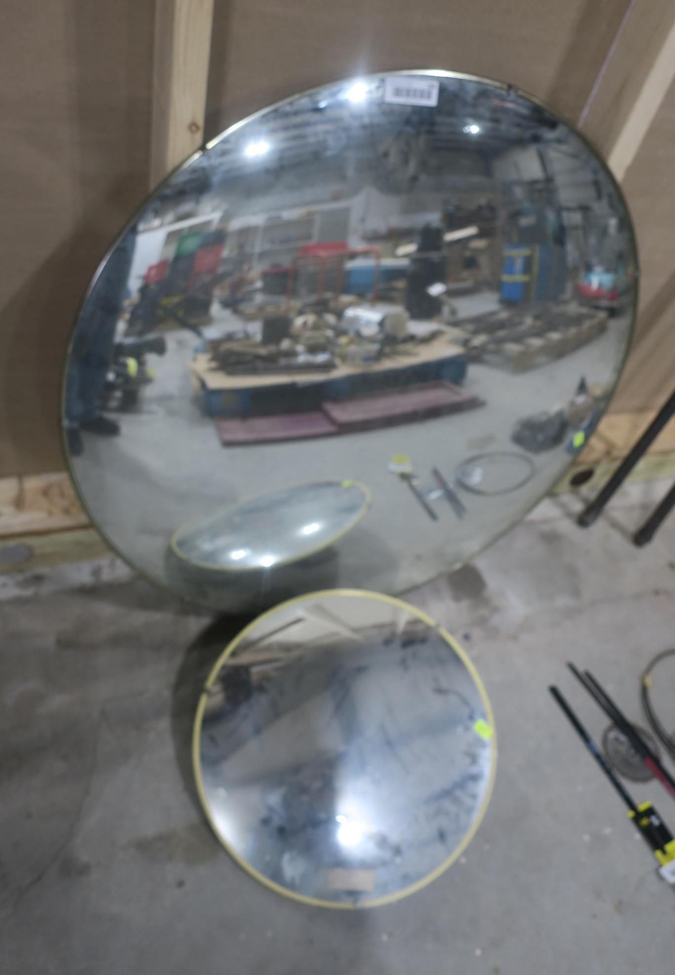 Two large convex mirrors leaning against a wall in a workshop or garage, reflecting shelves and tools.