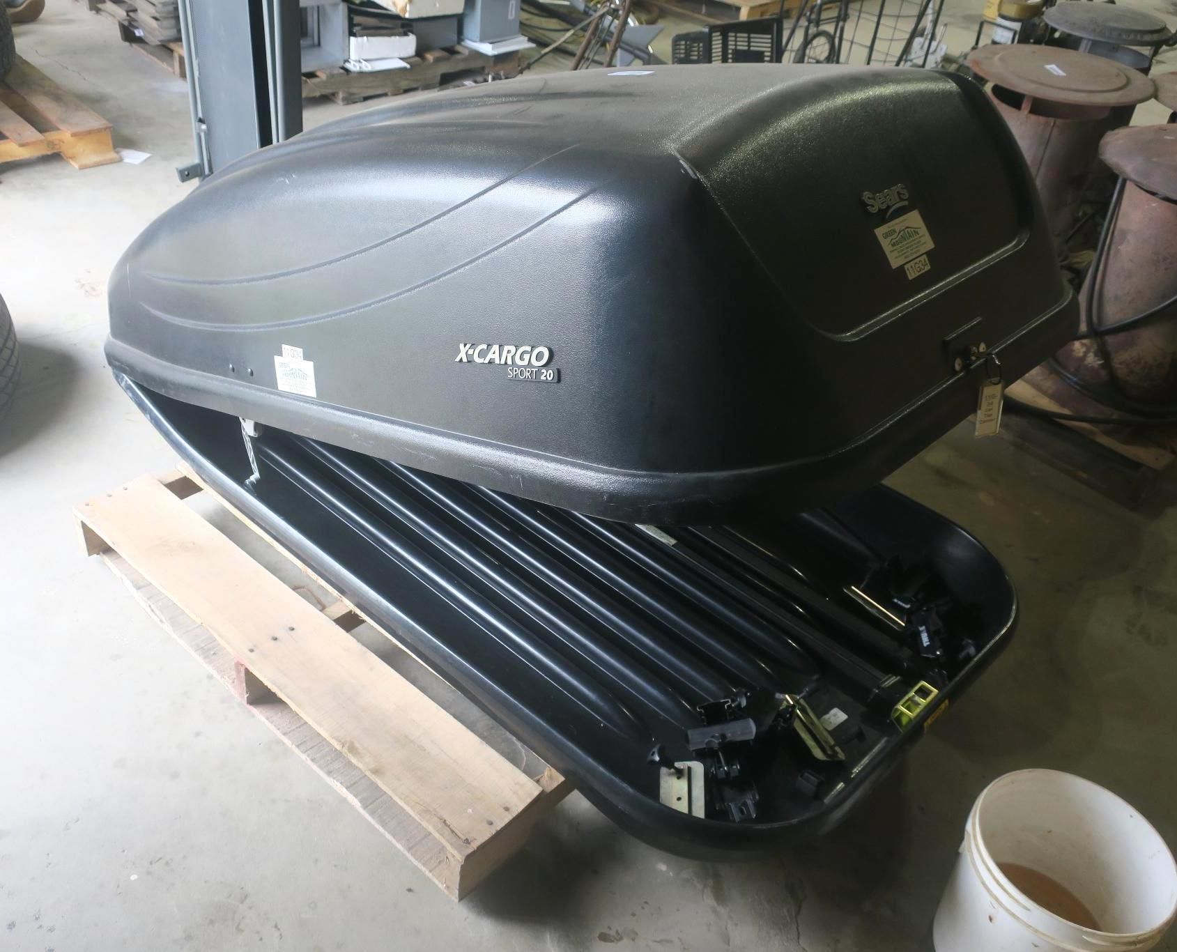Black hard-shell rooftop cargo box sitting on a wooden pallet in a garage or workshop.