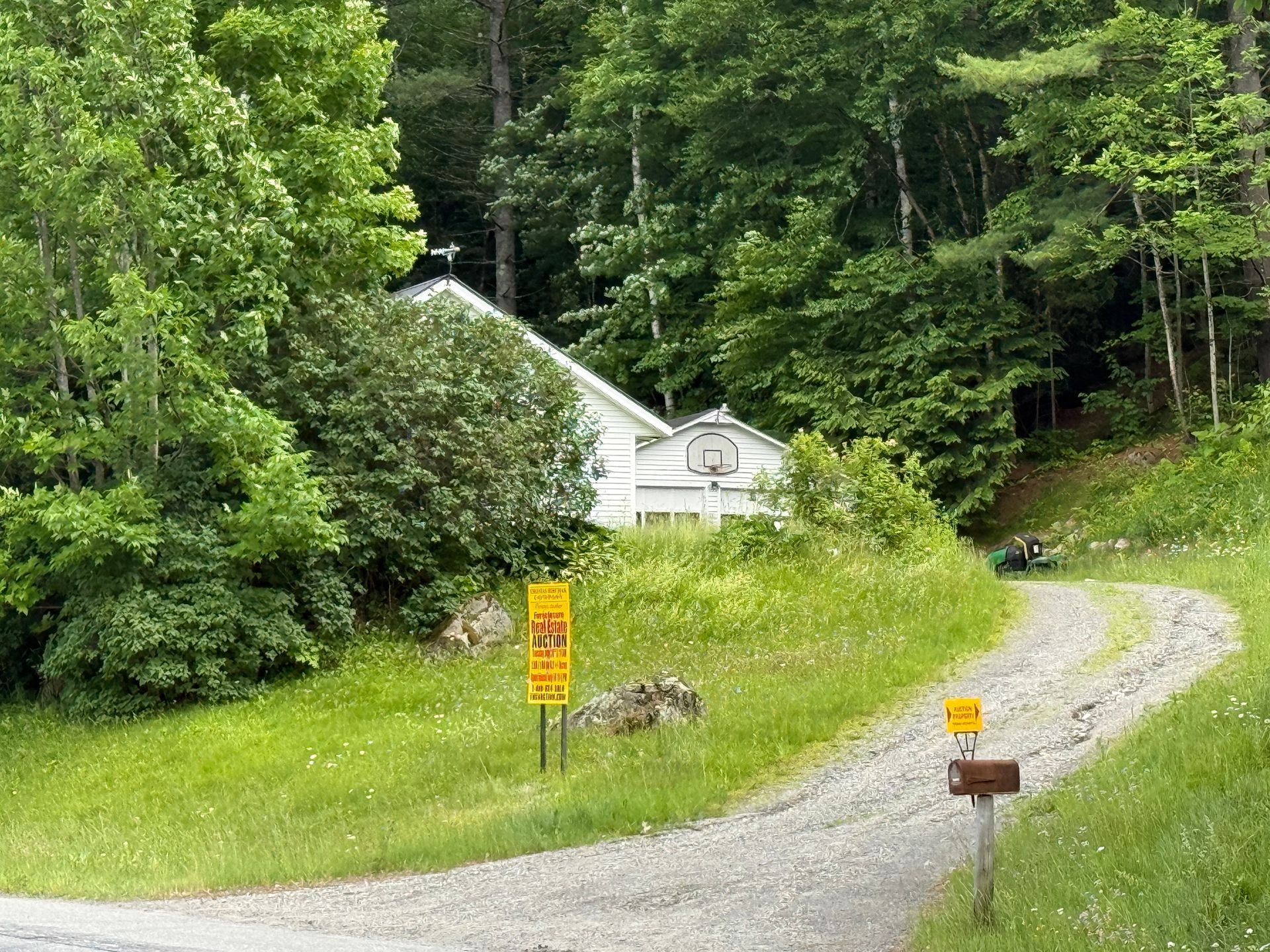 Gravel drive curving through a grassy wooded area toward a small white cabin with signs nearby.