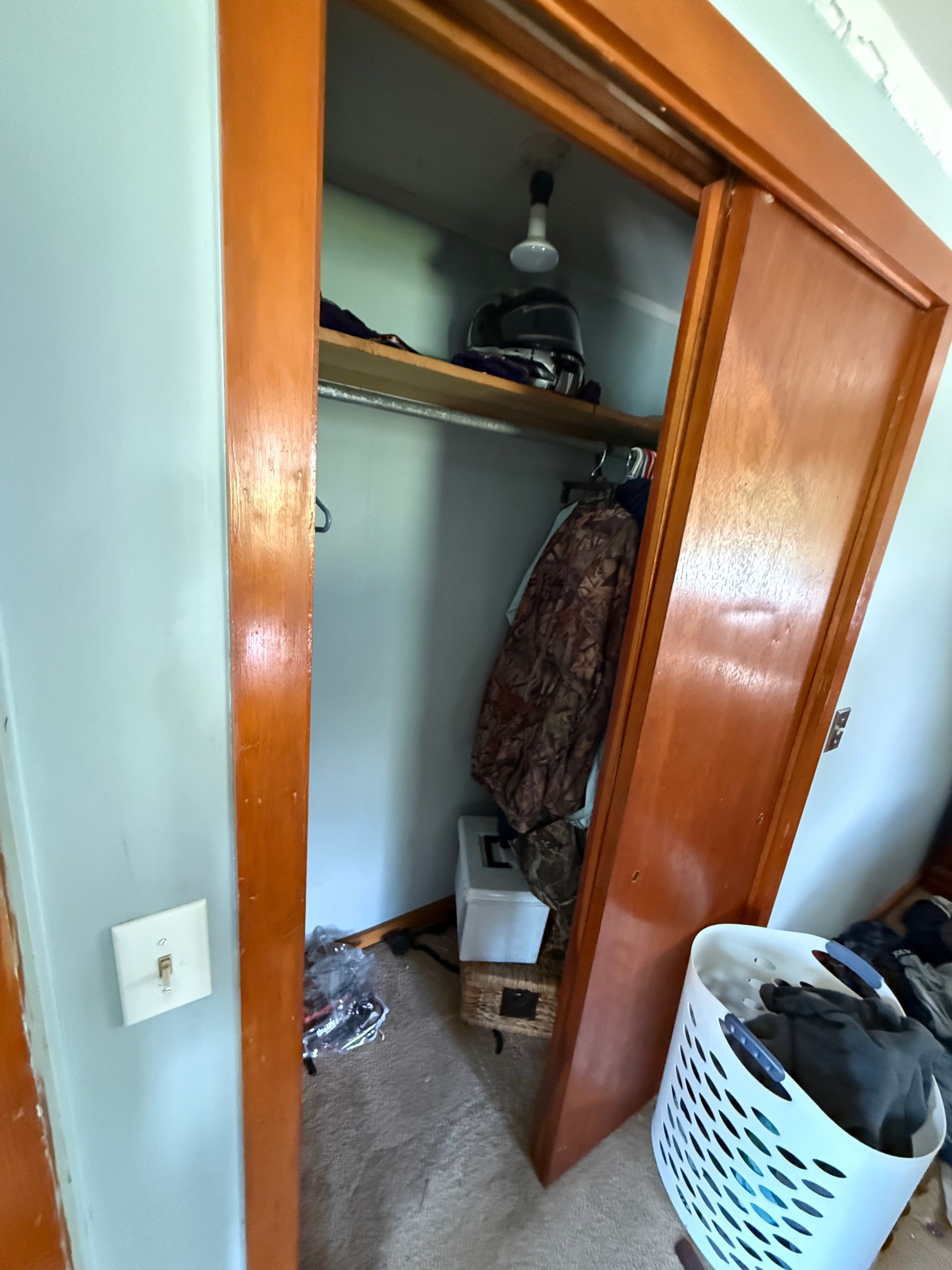 Open closet with hanging clothes, shelves, and a white laundry basket beside the door.