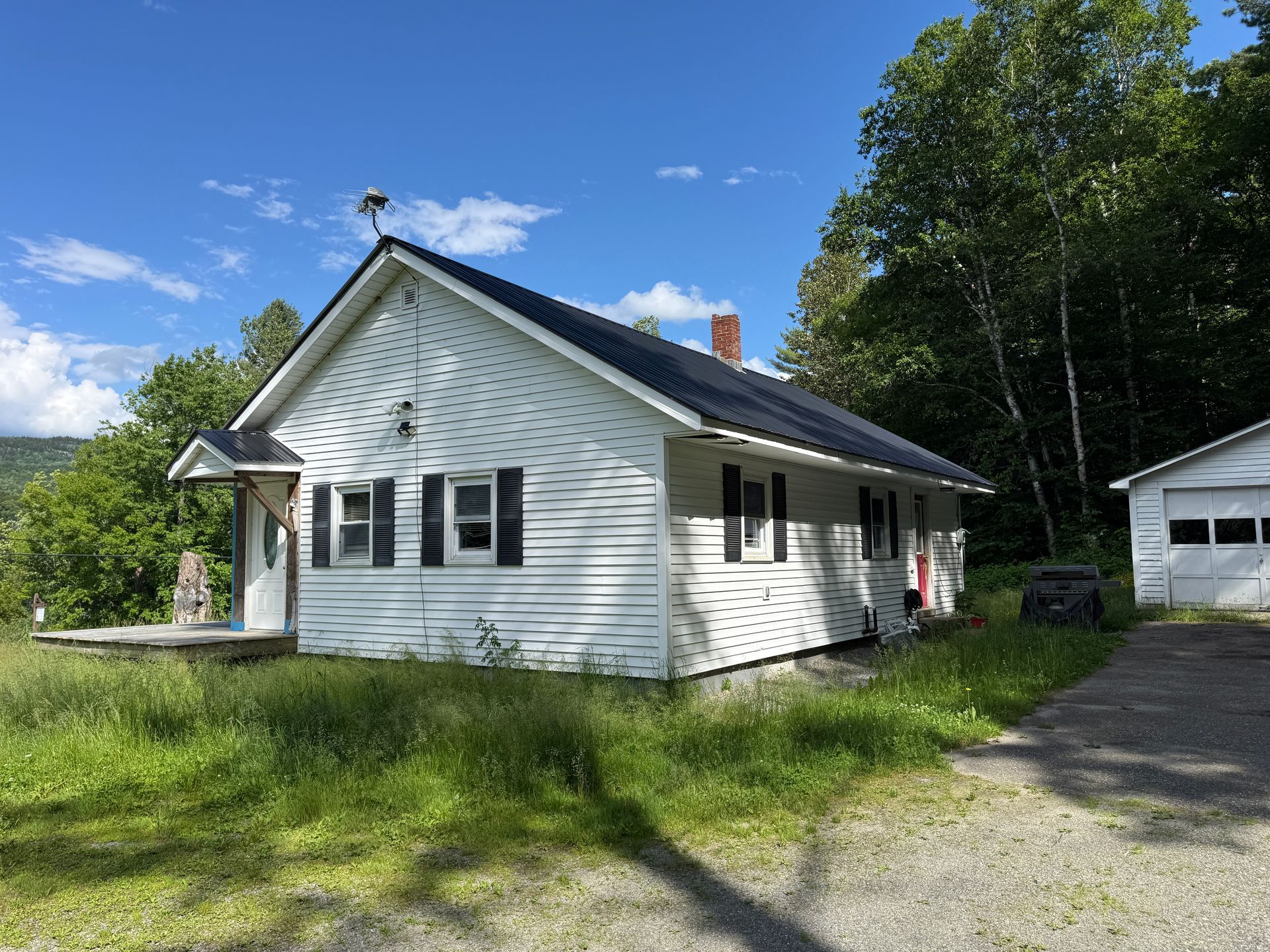 White house with dark roof on a sunny grassy lot, with a driveway and small outbuilding beside trees.