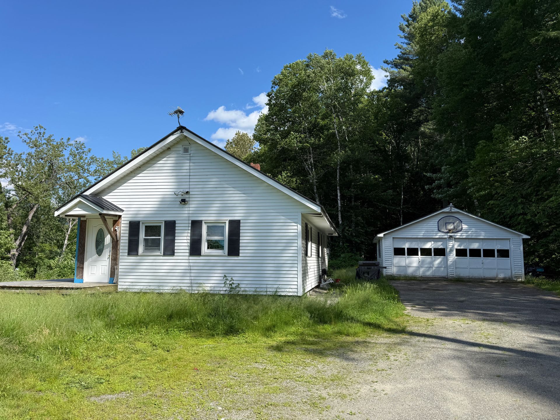 White house and detached garage with gravel driveway in a sunny, tree-lined yard