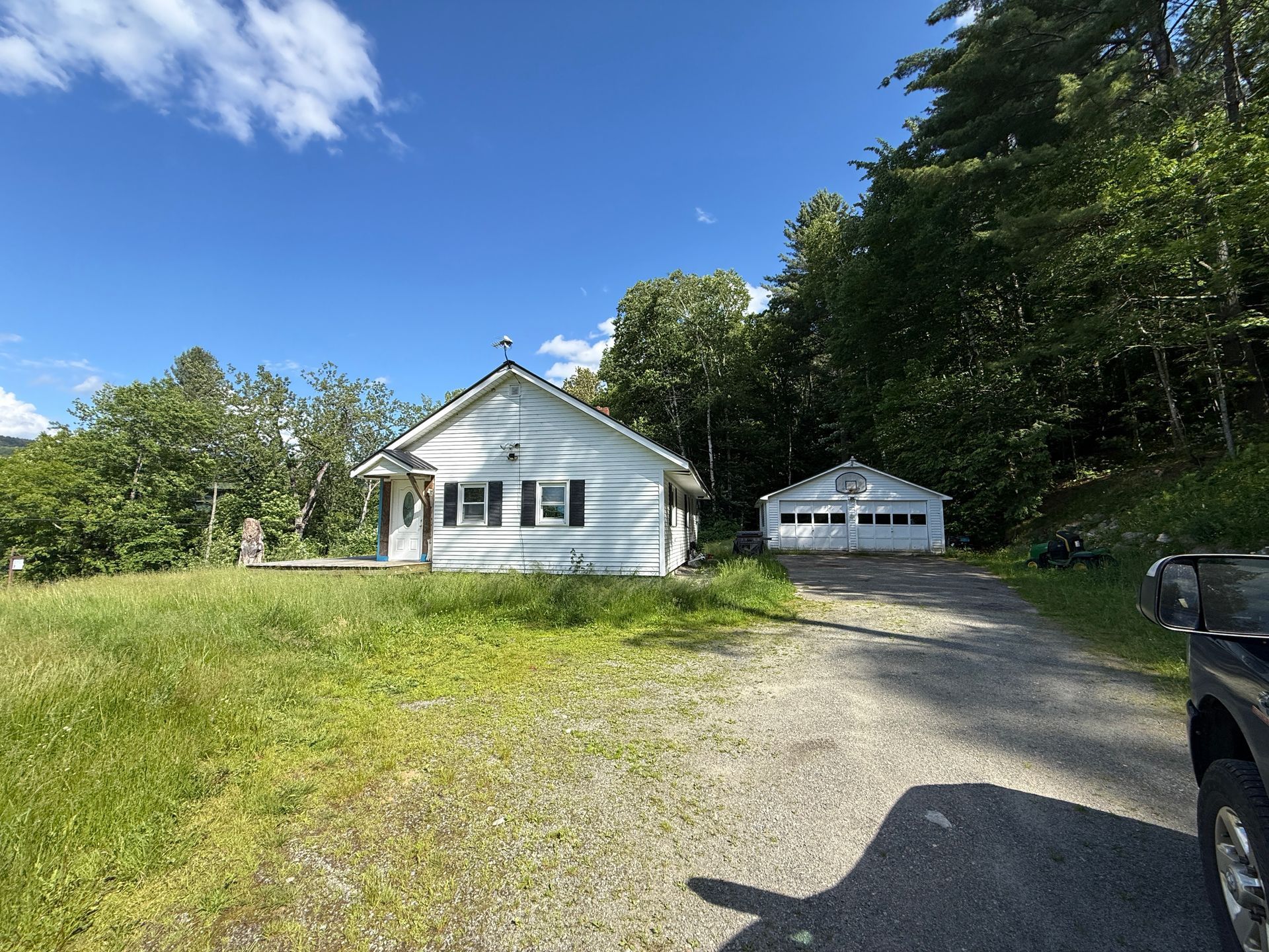 White house with gravel driveway, detached garage, and parked vehicle beside a wooded yard under a blue sky