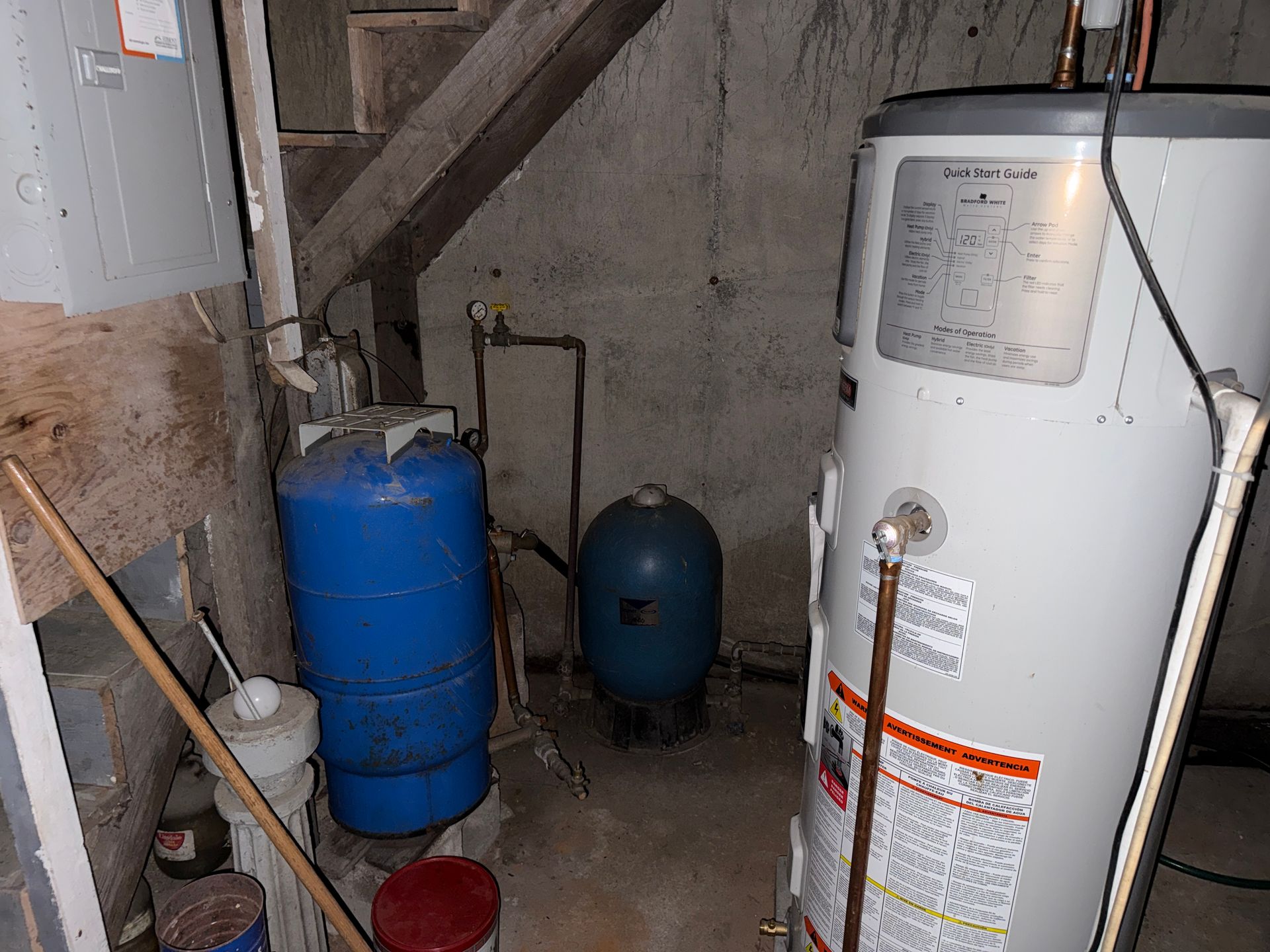 Basement utility area with water heater, blue tanks, pipes, and a concrete wall under stairs