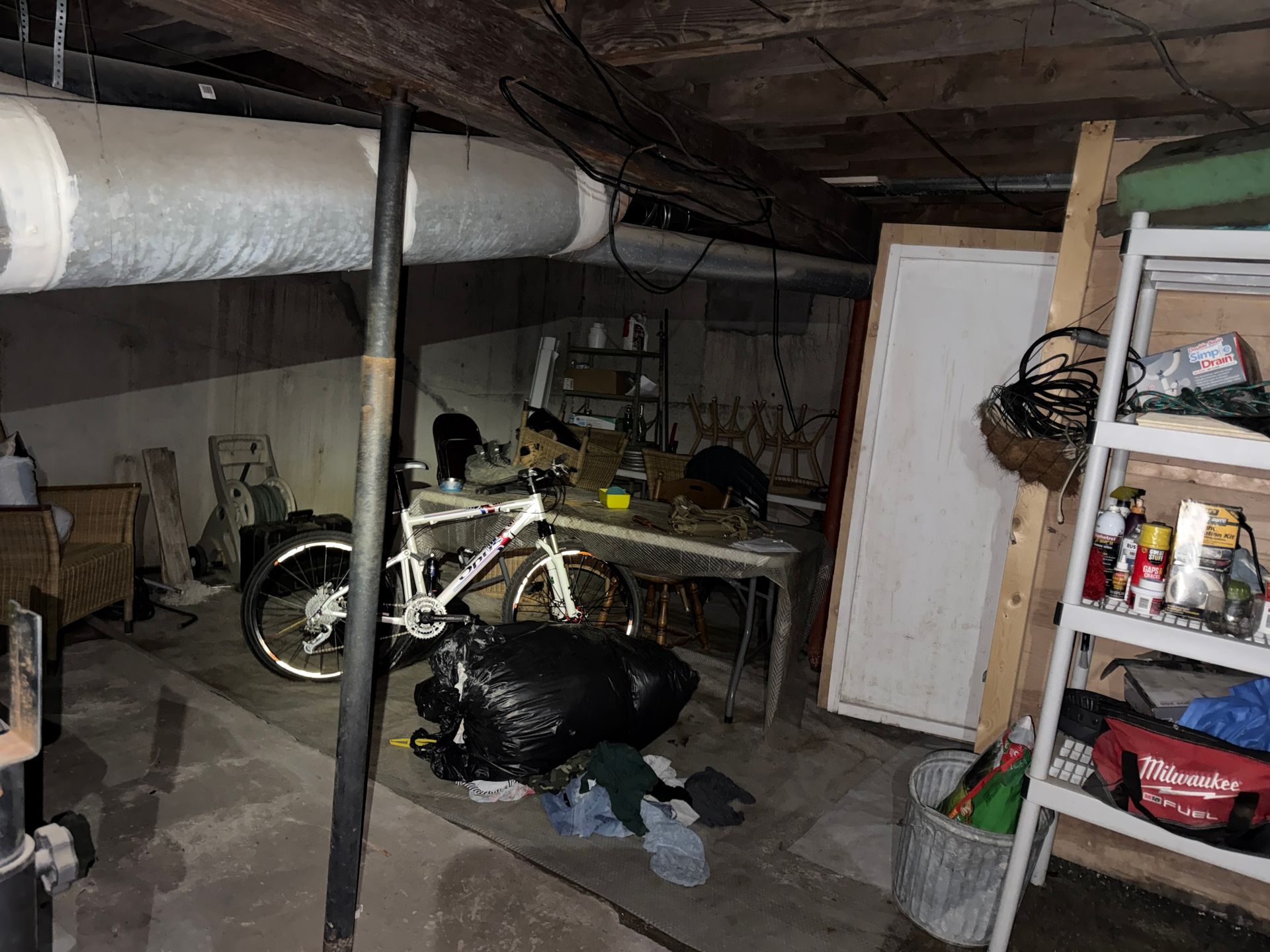 Basement with a bike, pipes, boxes, and shelves under low ceiling.