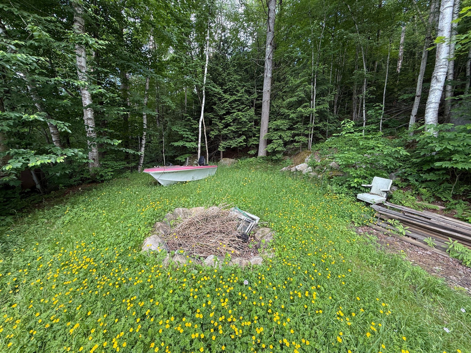 Backyard fire pit surrounded by yellow flowers and trees, with a red canoe and white chair nearby