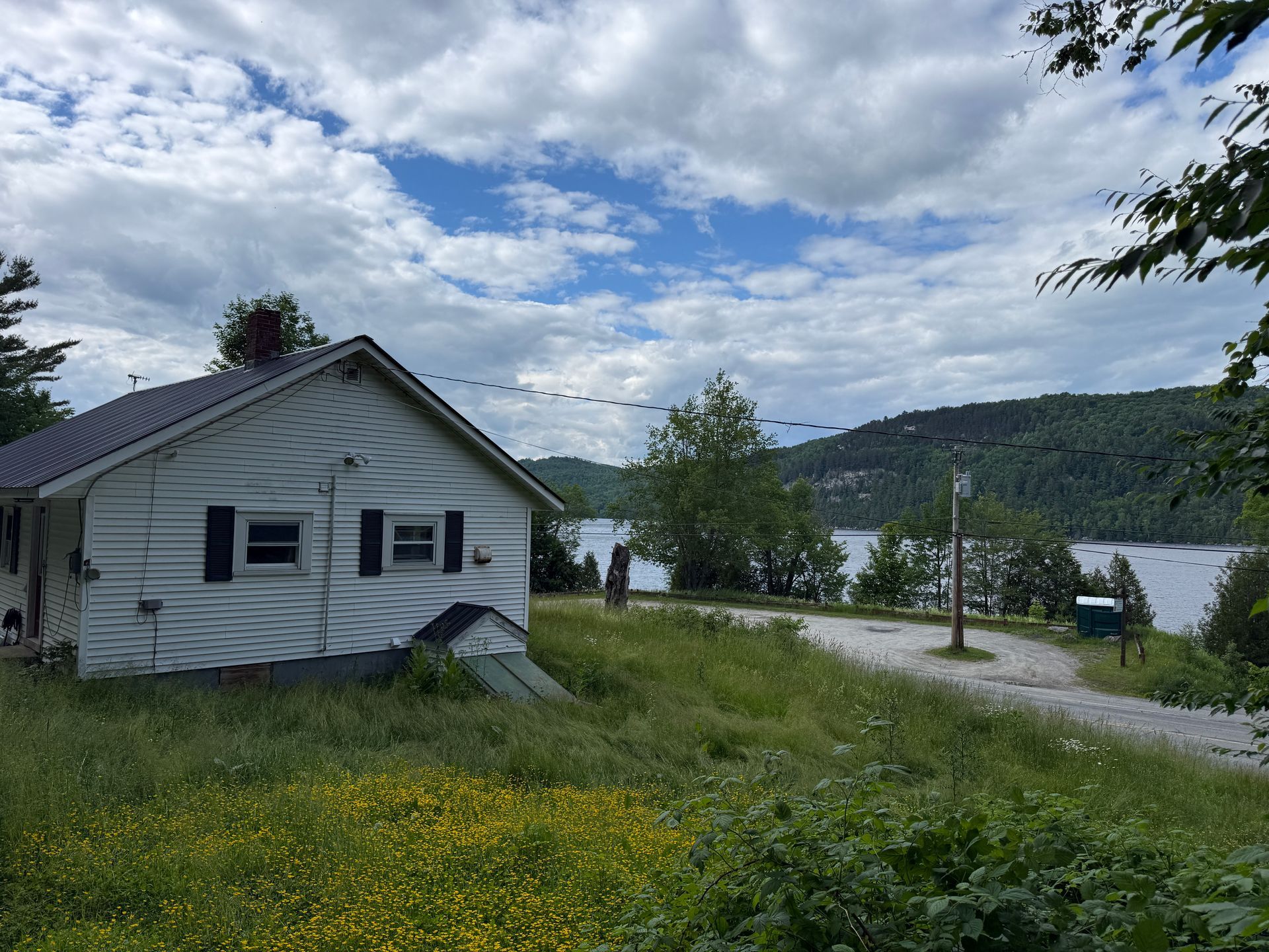 White lakeside cabin under cloudy sky with grassy yard, gravel road, and water in the background