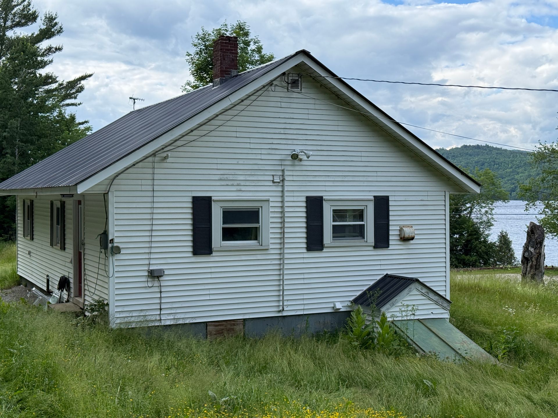 Small white house with dark shutters, set on a grassy hill near water and trees.