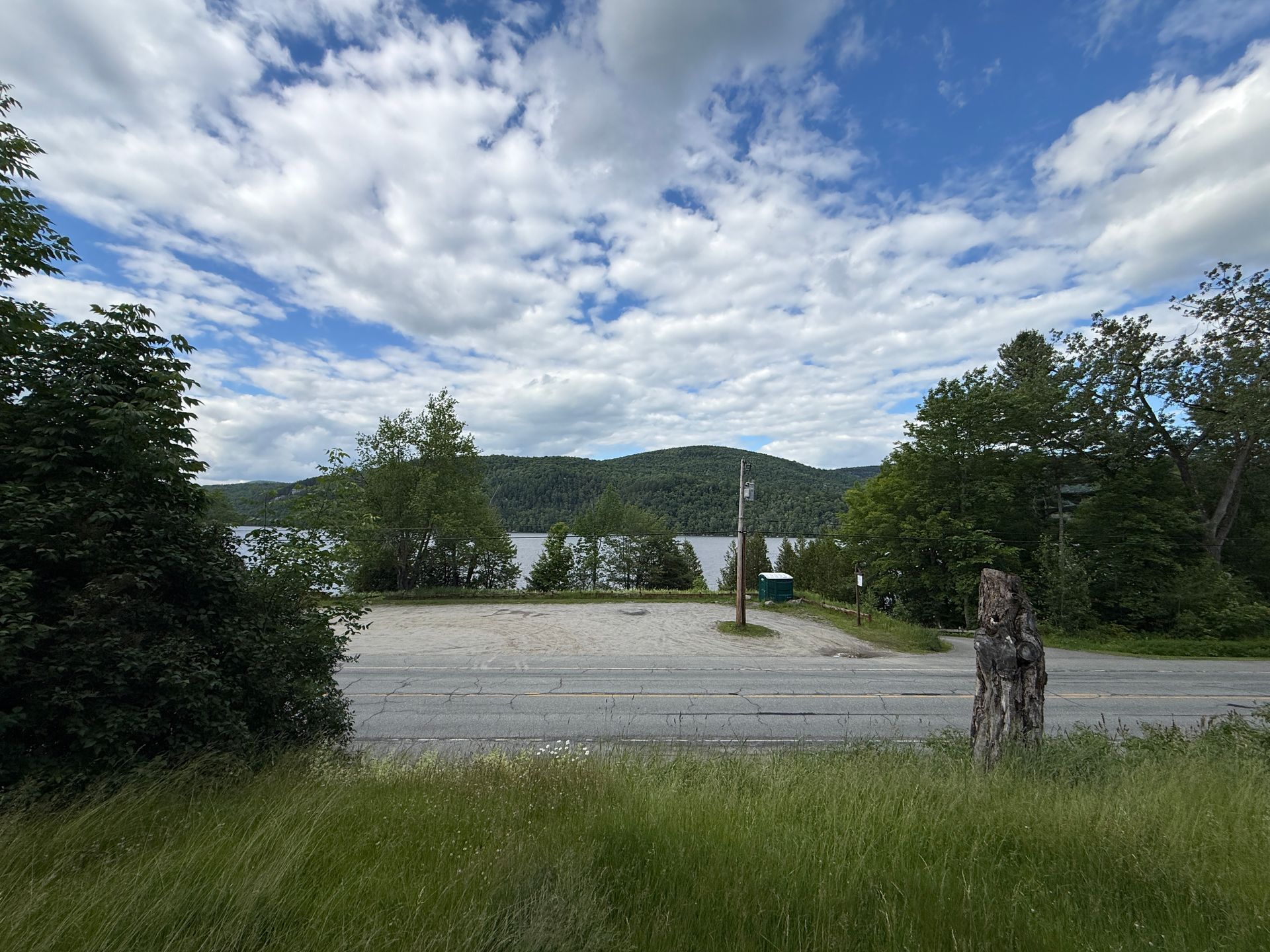 Lakeside overlook with gravel lot, trees, hills, and cloudy sky