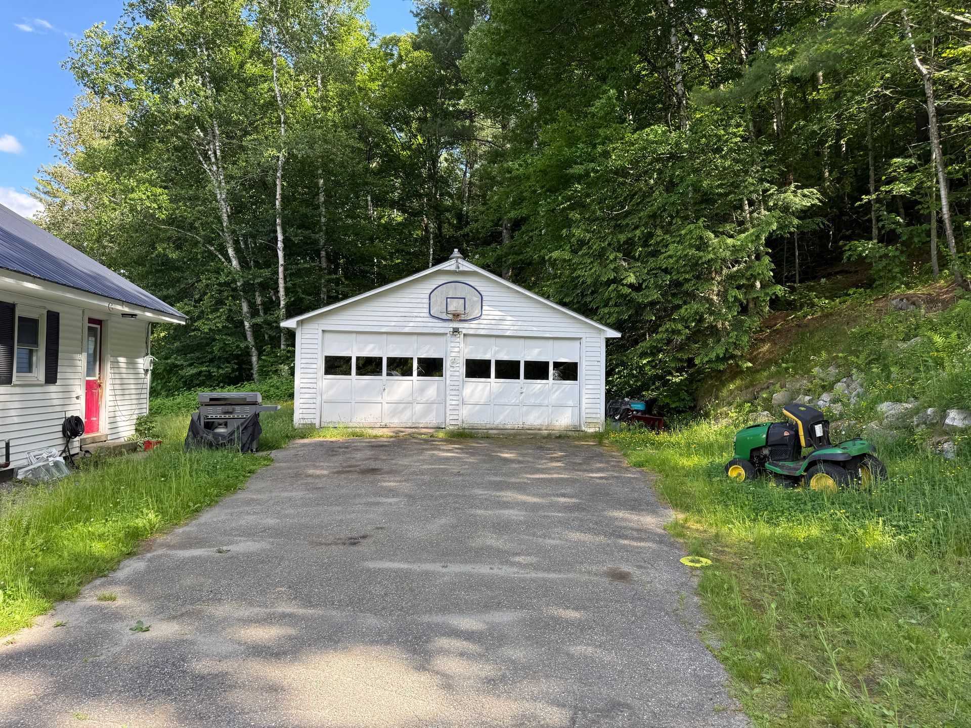 Gravel driveway leading to a white garage with parked lawn tractor, surrounded by trees and grass