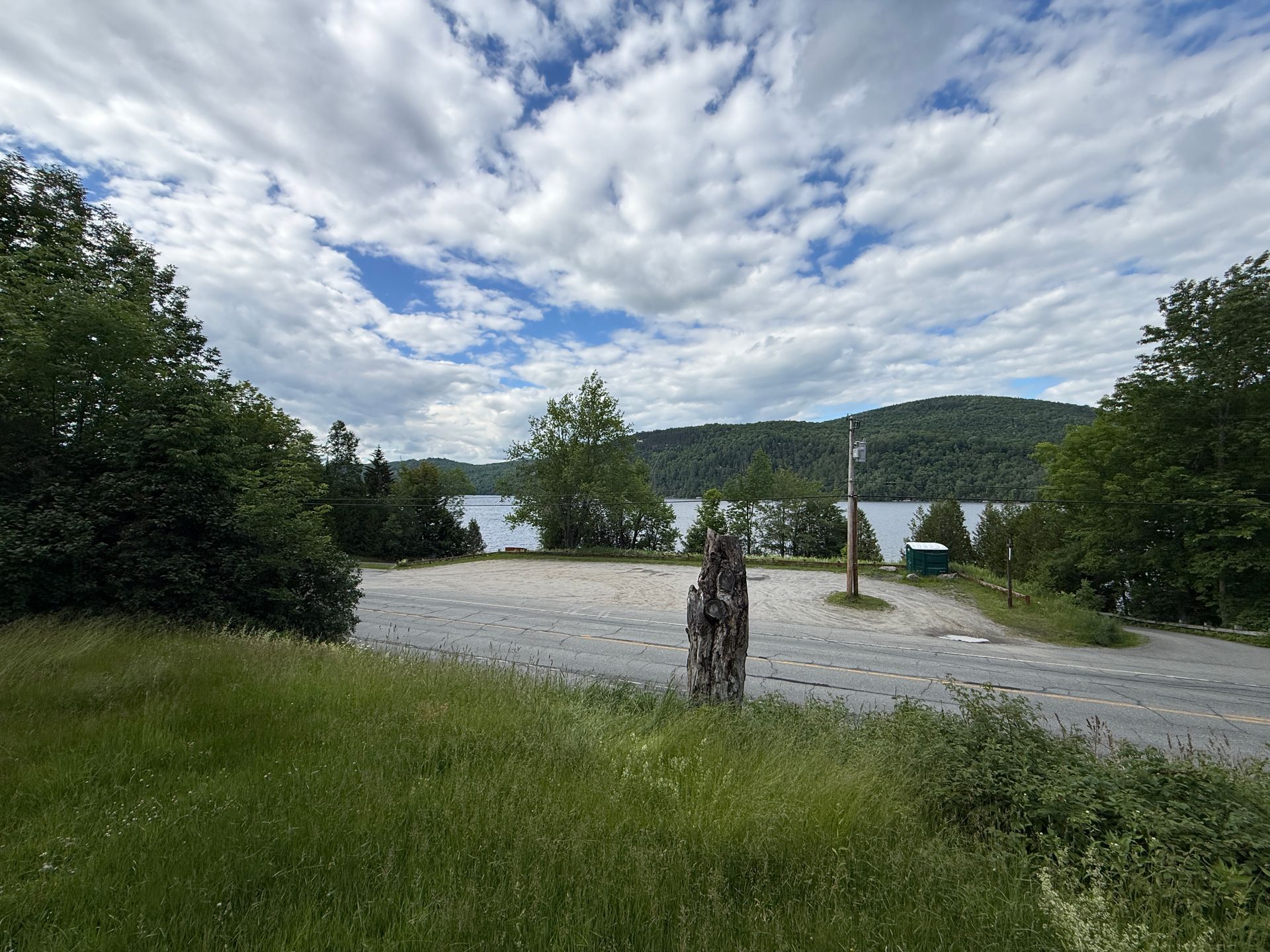 Lakeside gravel area with trees, hills, and a cloudy sky