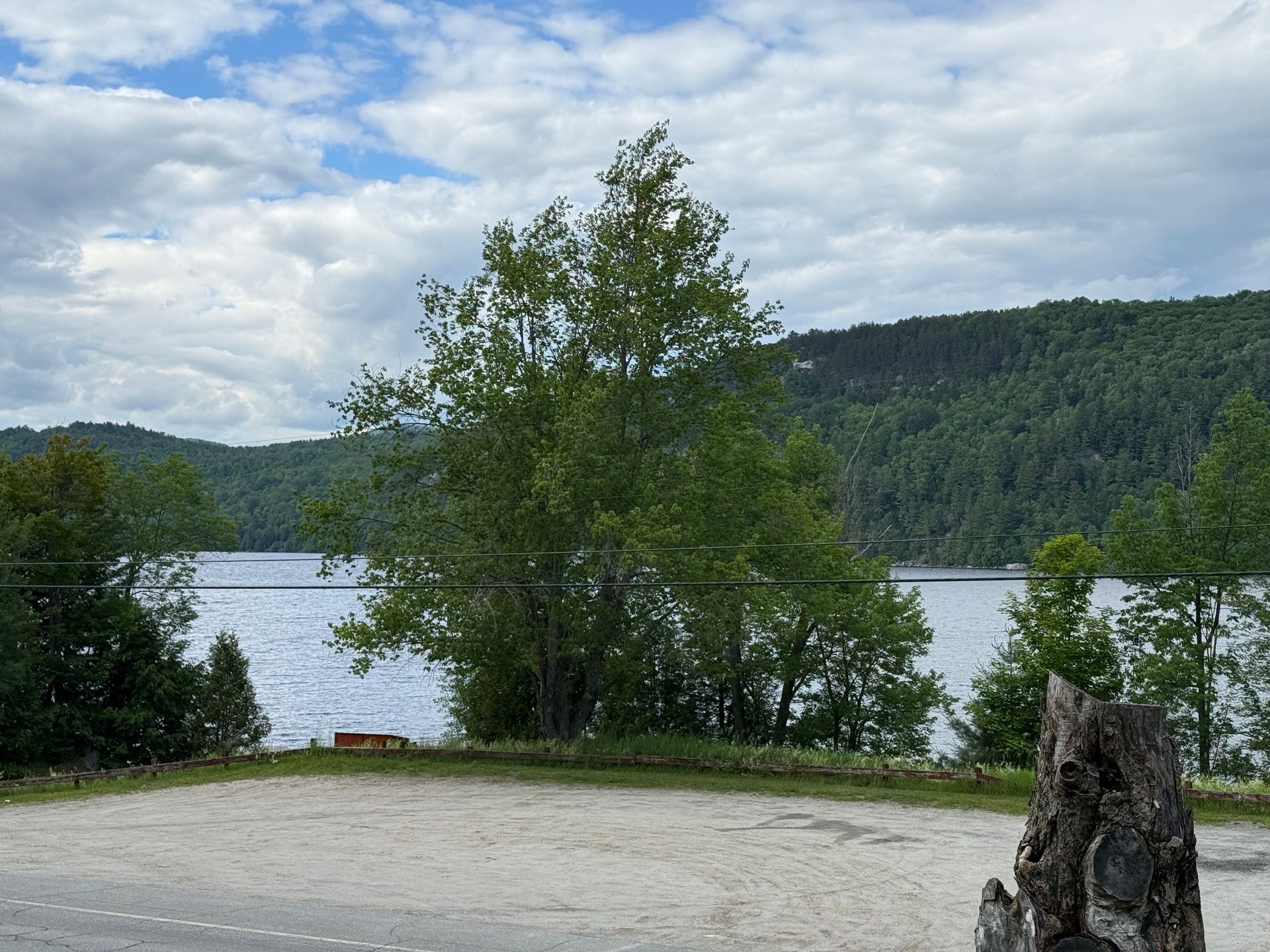 Lakeside view with trees, a sandy shore, and forested hills under a cloudy sky