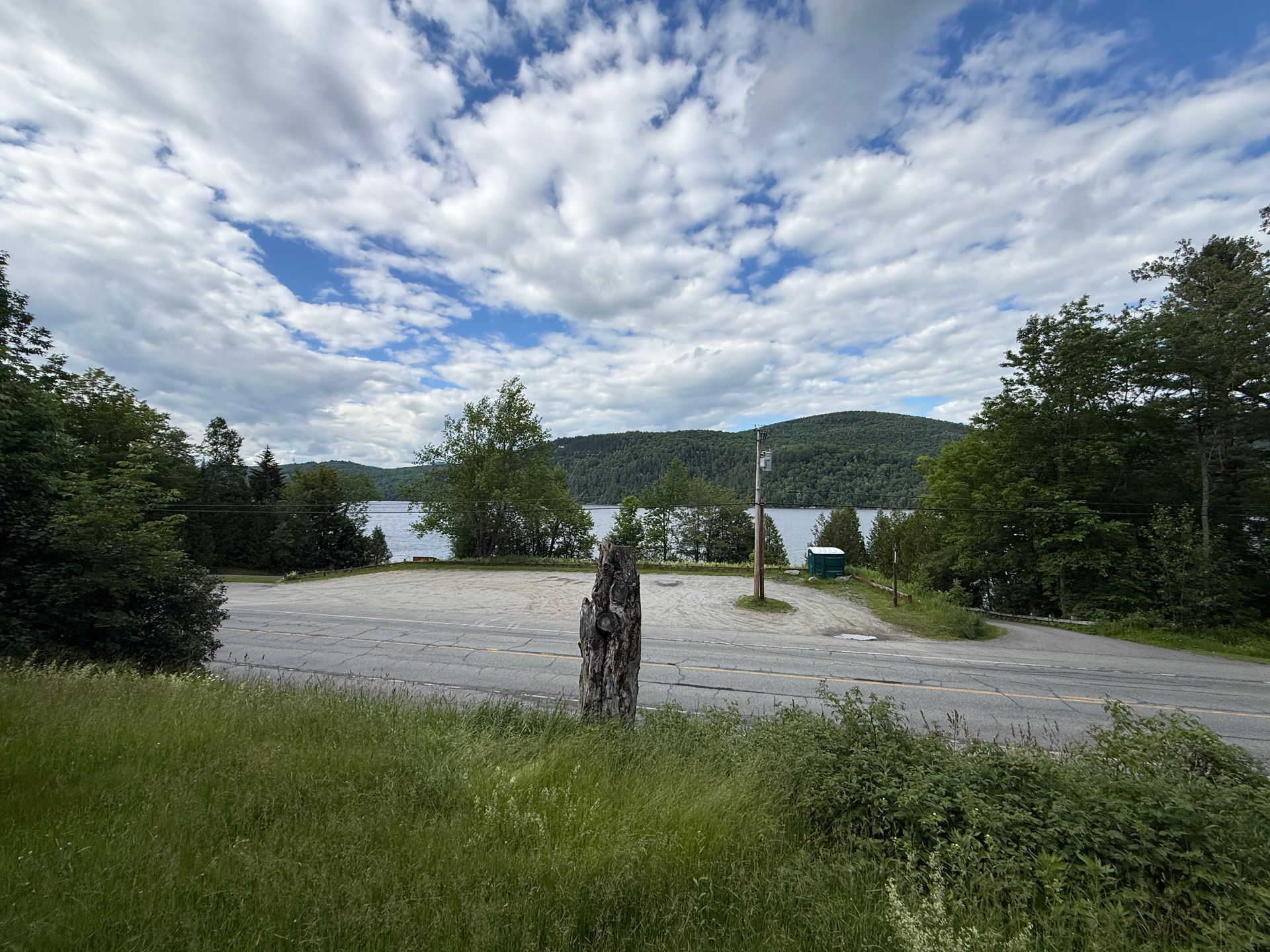 Gravel lakeside parking area with trees, a stump, and cloudy sky over distant hills