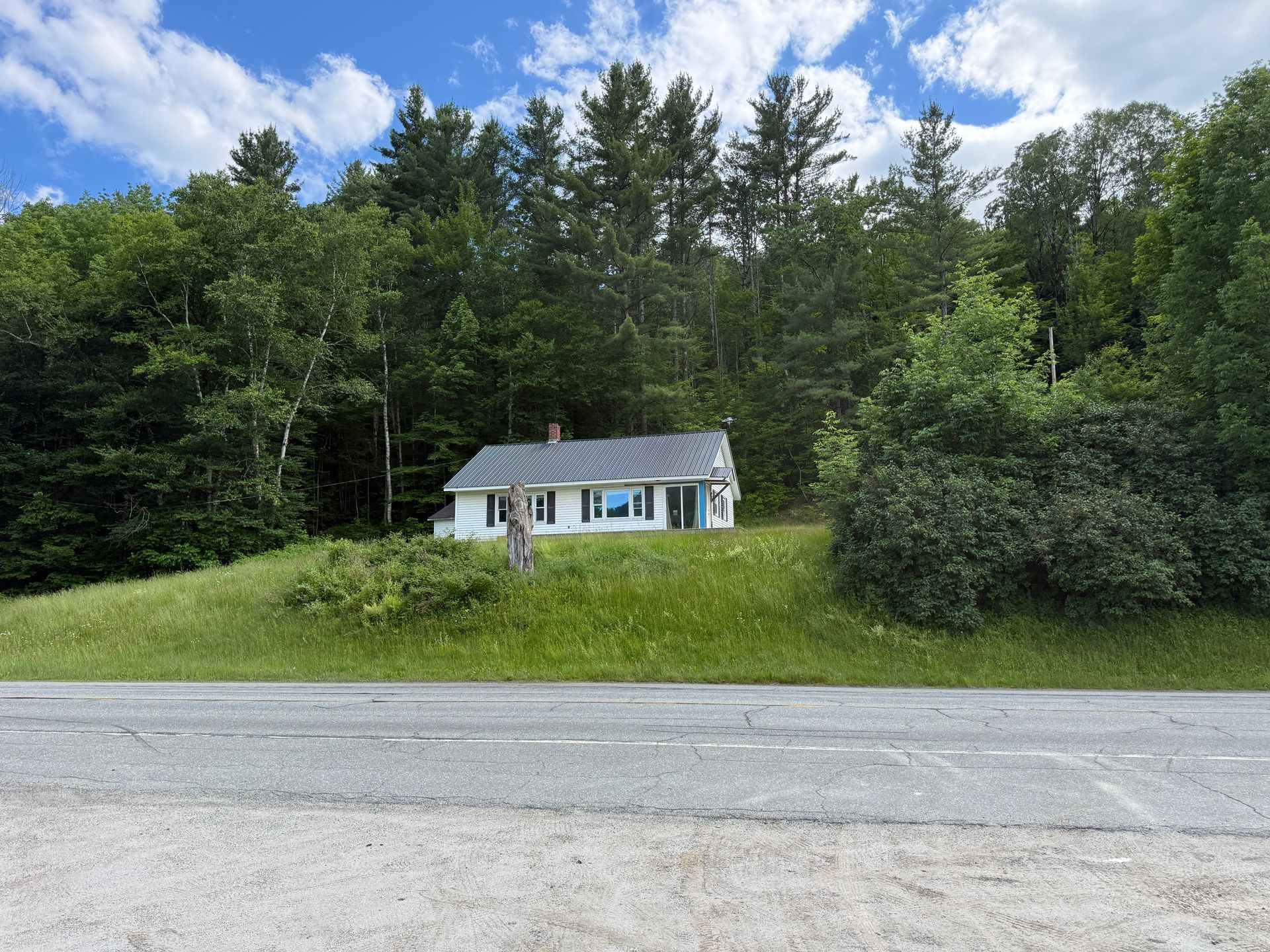 Small white house with blue shutters beside a gravel road, set against a green forest under a cloudy sky