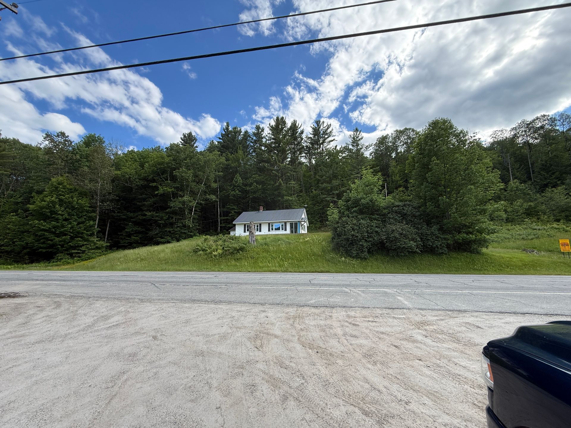 Small white house across a gravel road, surrounded by green trees under a cloudy blue sky
