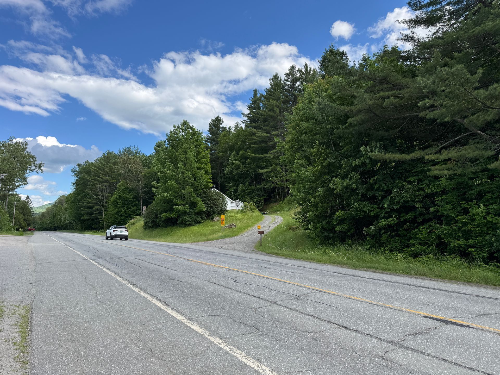 Two-lane road curving past dense green trees under a blue sky with clouds, with one car approaching