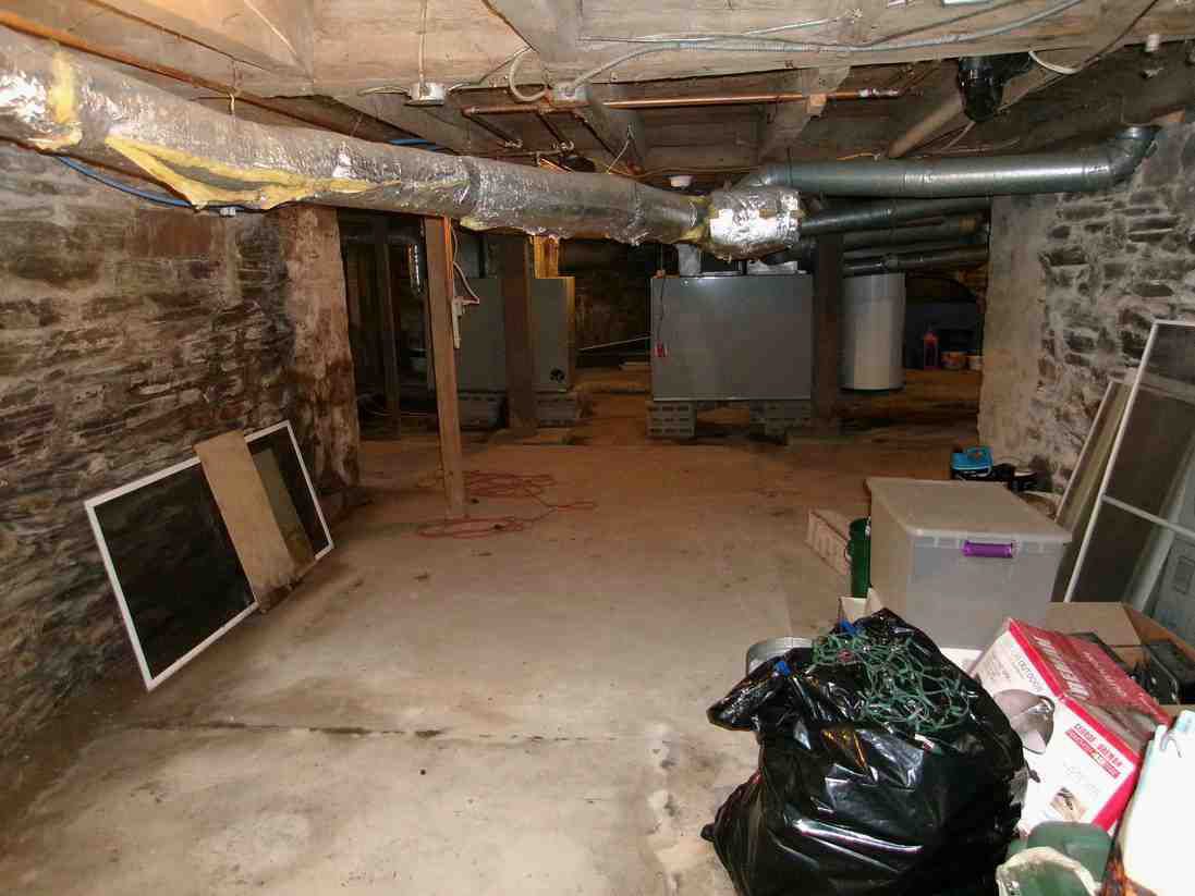 Unfinished basement with stone walls, exposed pipes, a furnace, and stored boards and bags on the floor.