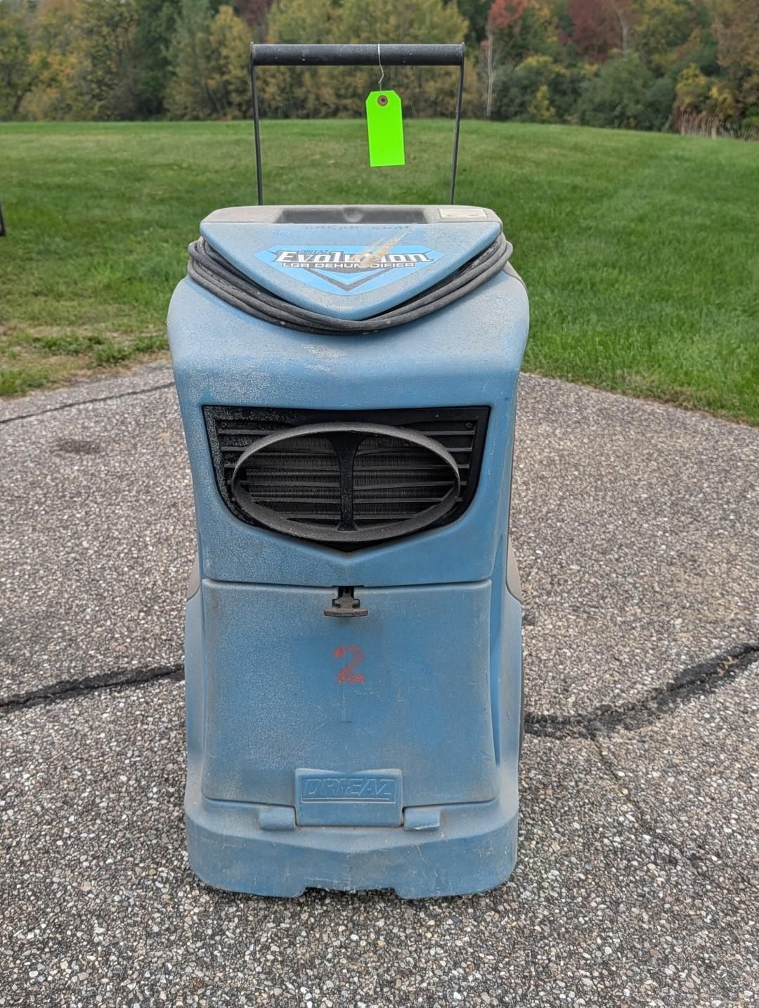 Blue wheeled trash bin on a paved path with a bright green tag and black oval front vent.