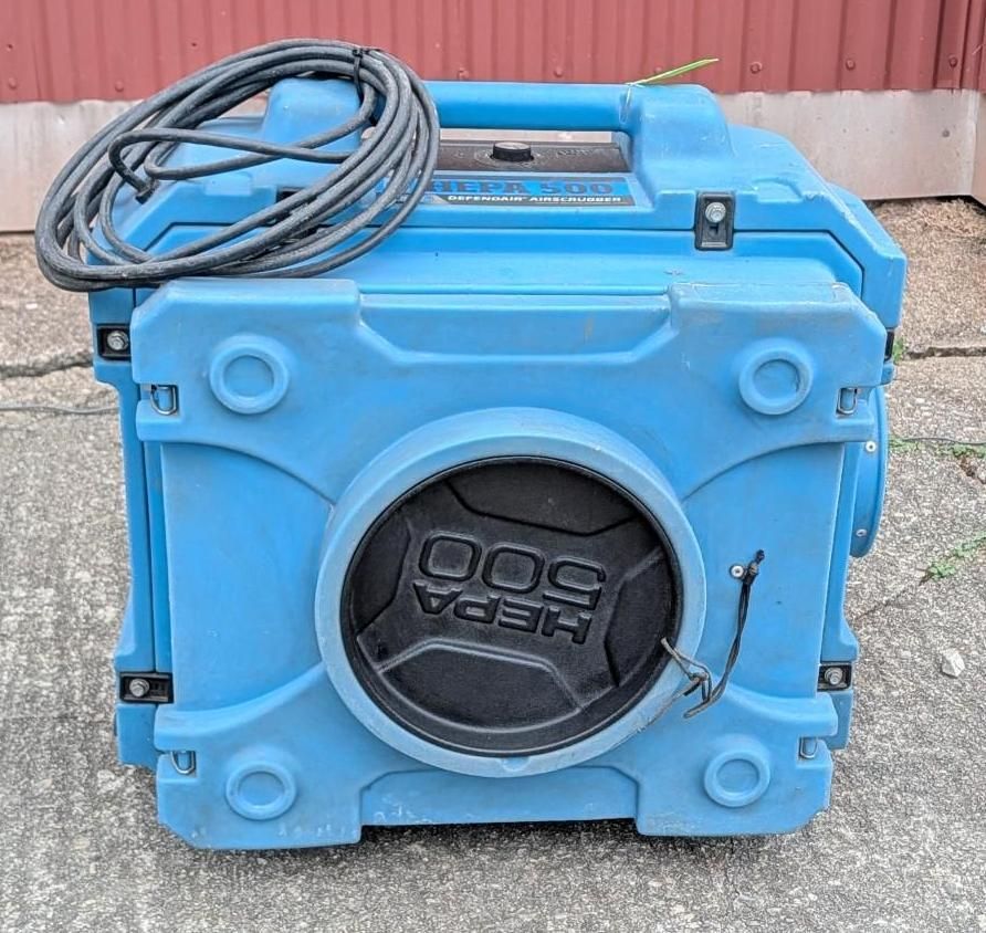 Blue industrial machine with coiled black hose, sitting outdoors on gravel against a red wall