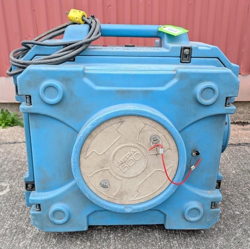 Blue portable plastic tank with a round beige hatch and coiled cables on top, against a red wall.