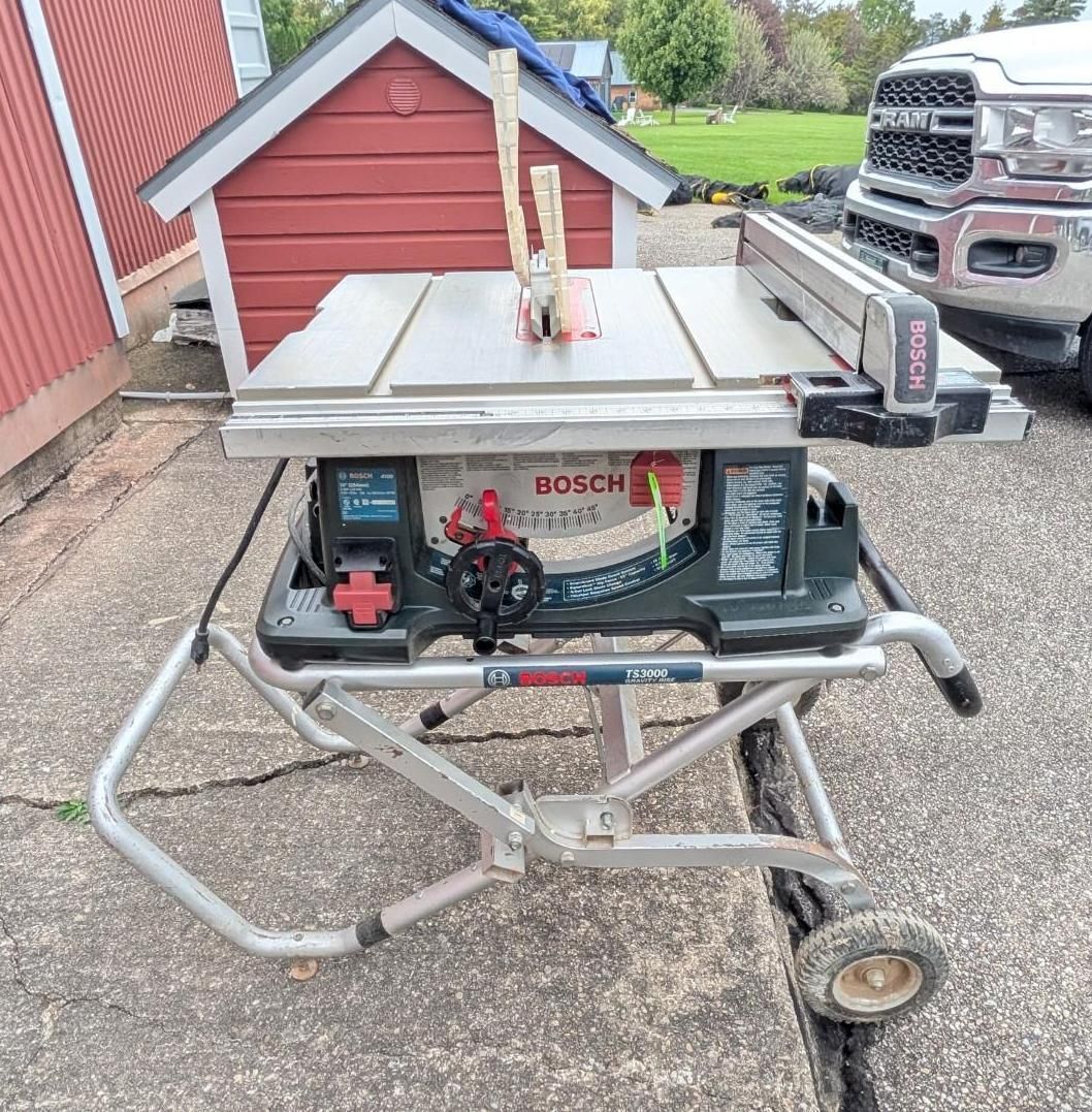 Portable Bosch table saw on a wheeled stand outdoors beside a red shed and pickup truck.