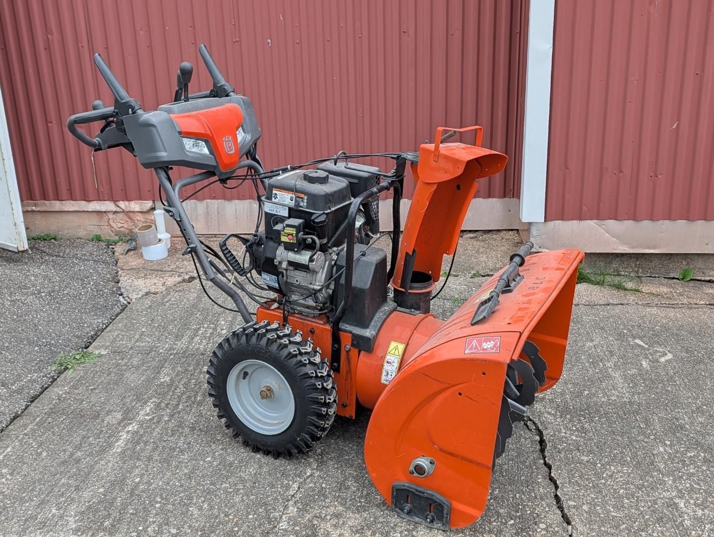 Orange snow blower parked on gravel beside a red building