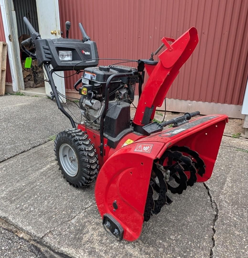 Red snow blower with red chute and black wheels parked on a driveway beside a red building.