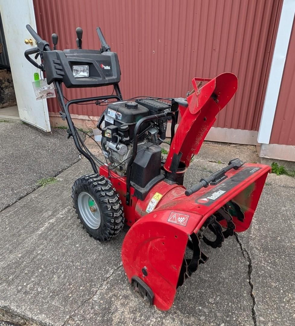 Red snow blower with black handlebars parked on a concrete driveway in front of a red building.