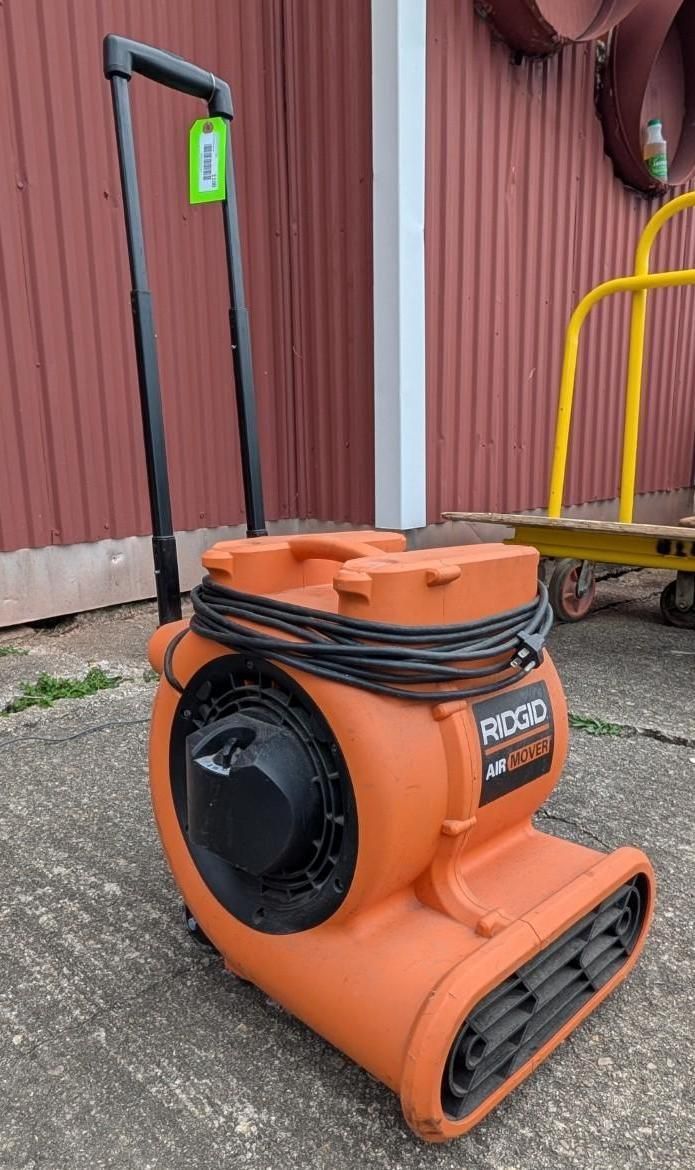 Orange industrial blower on a gravel surface beside a red building, with a black handle and wheels.