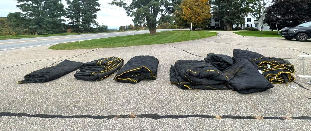 Black moving blankets laid out in rows on a paved roadside with trees and parked vehicles in the background
