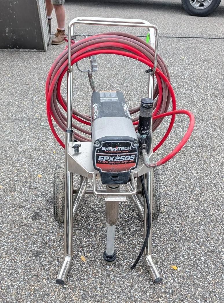 Portable airless paint sprayer with red hose on a metal cart, parked on asphalt outdoors