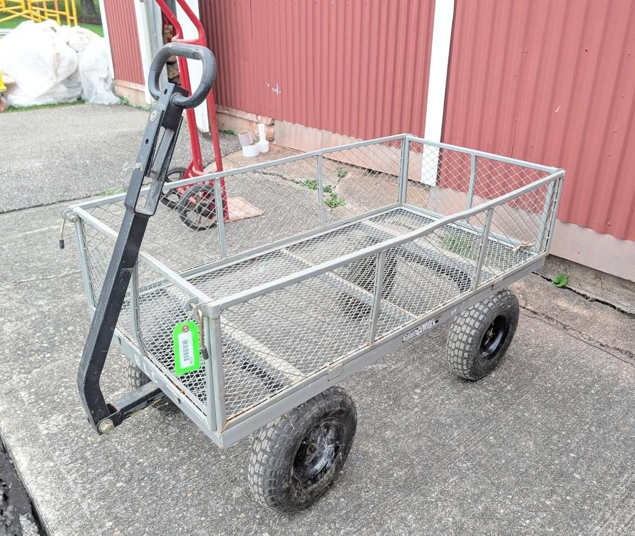 Black hand truck towing a wire garden cart with four wheels on pavement beside a red wall