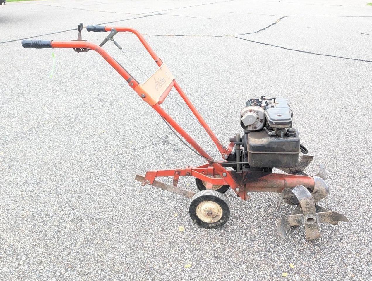 Red tiller on pavement, with black engine, orange handle, and metal tines.