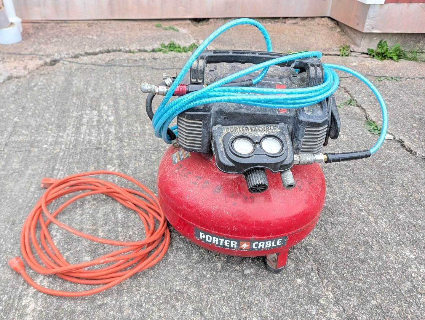 Red air compressor with coiled blue hose and orange extension cord on concrete outdoors