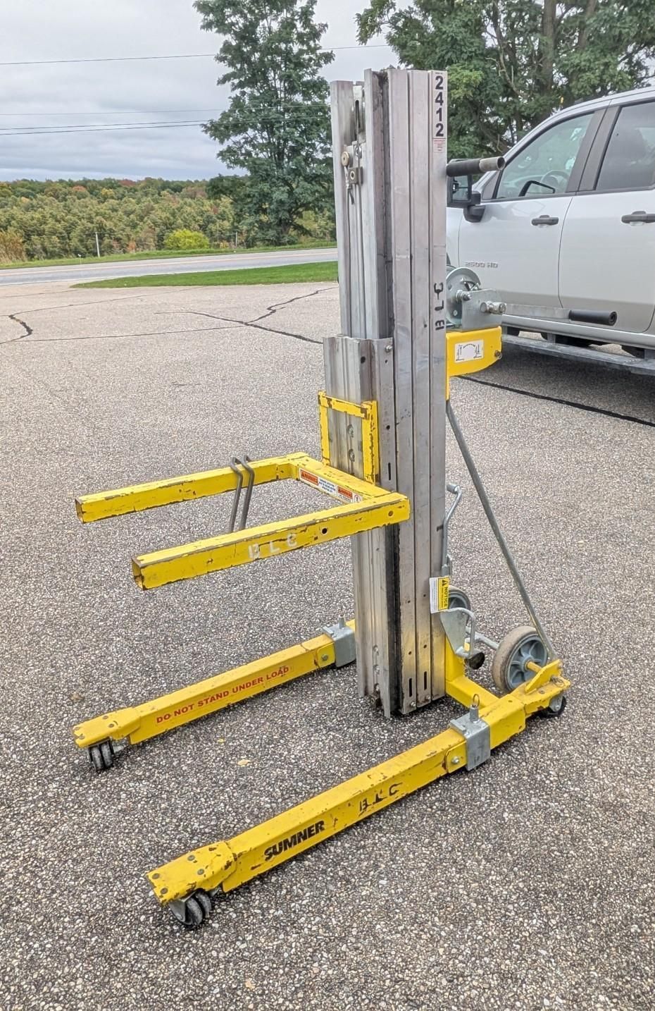 Yellow forklift mast with four prongs beside a silver truck on pavement outdoors