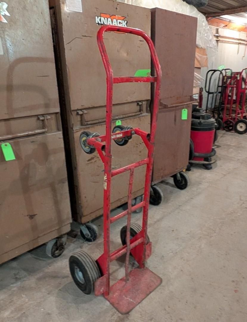 Red hand truck standing in a warehouse aisle beside large brown carts or boxes.