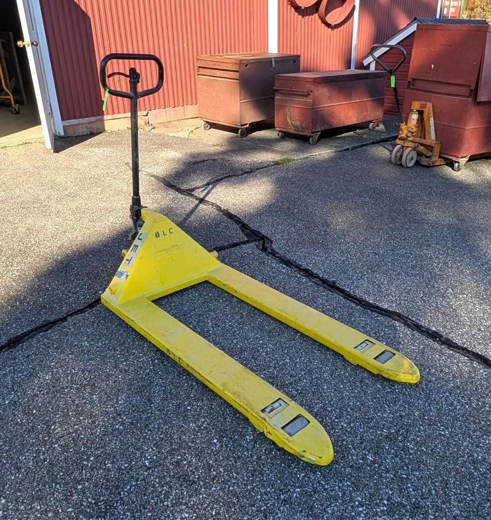 Yellow pallet jack on gravel in front of red industrial building.