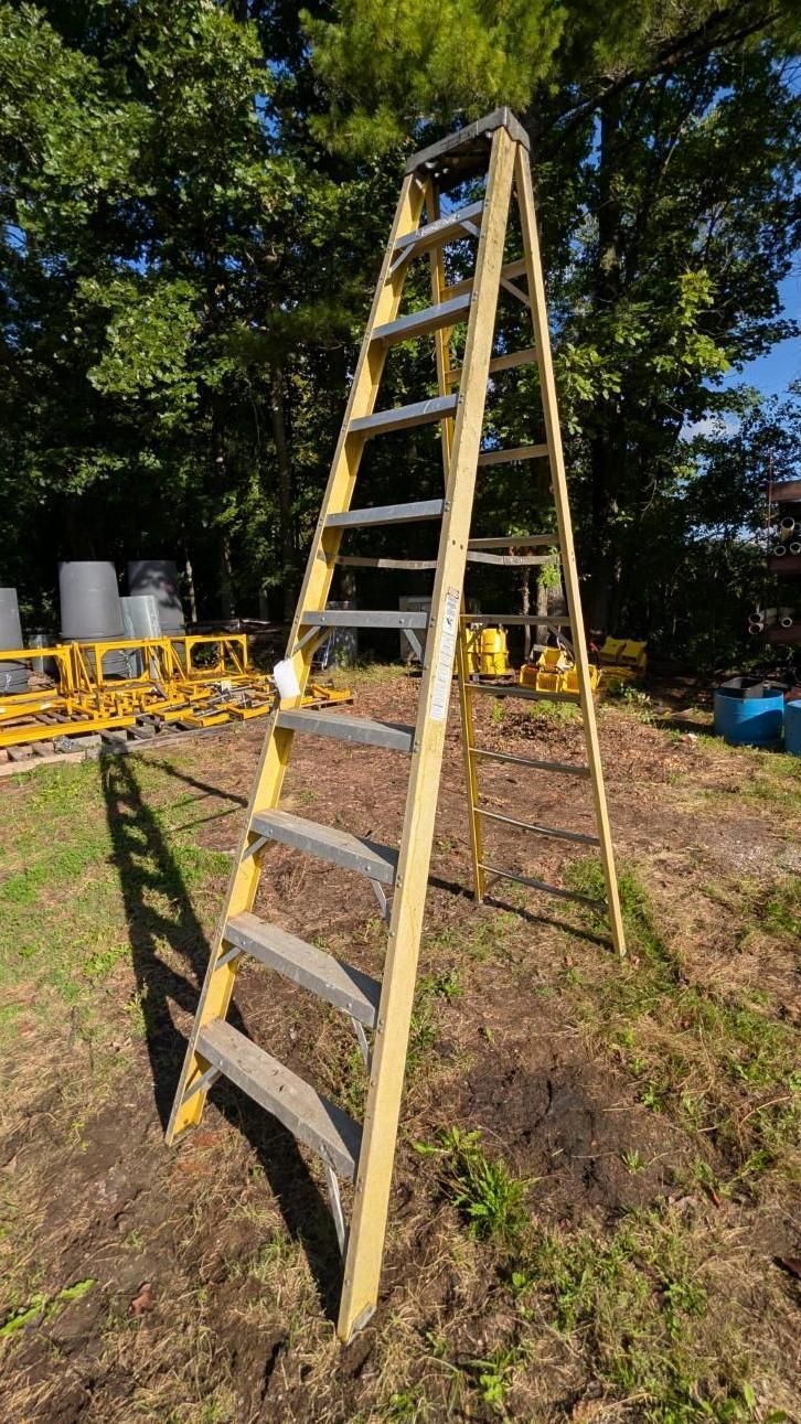Yellow A-frame ladder standing on grass outdoors near trees and construction barriers.