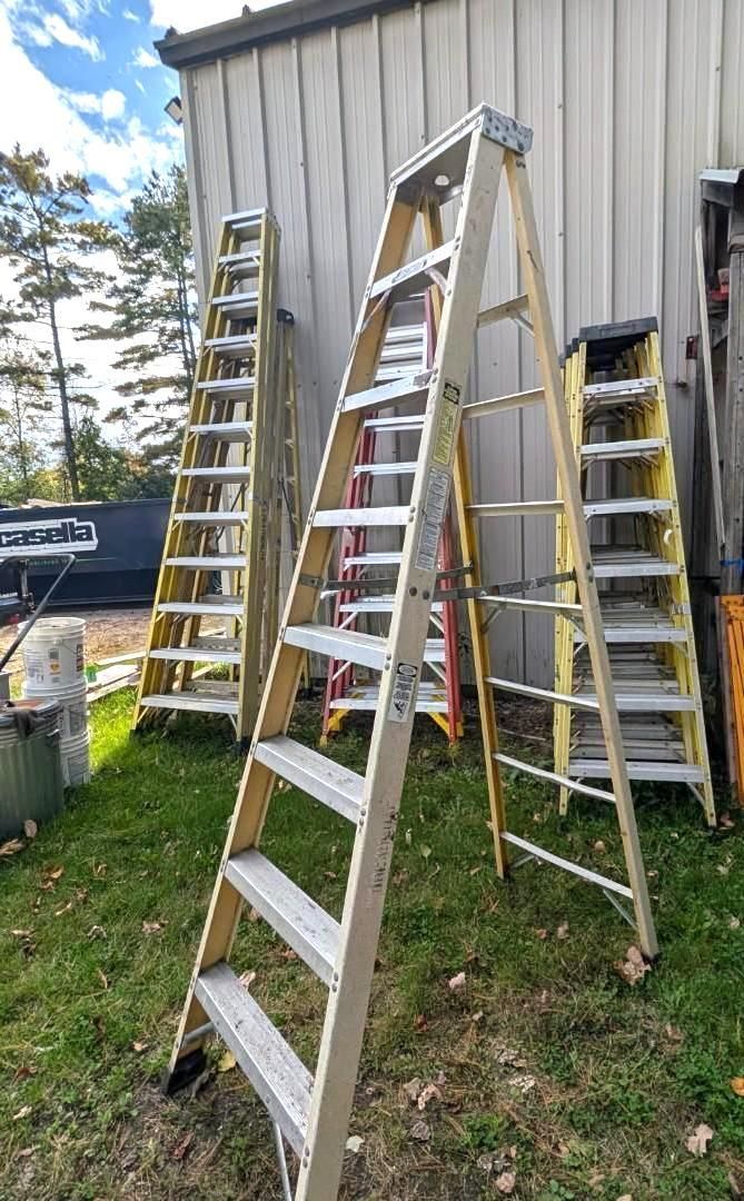 Several ladders lean against a metal-sided shed in a grassy yard under a partly cloudy sky