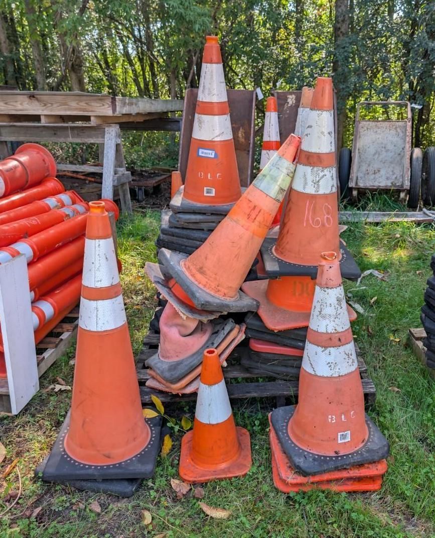 Stack of orange and white traffic cones piled outdoors on grass, with a wooden fence and trees behind.