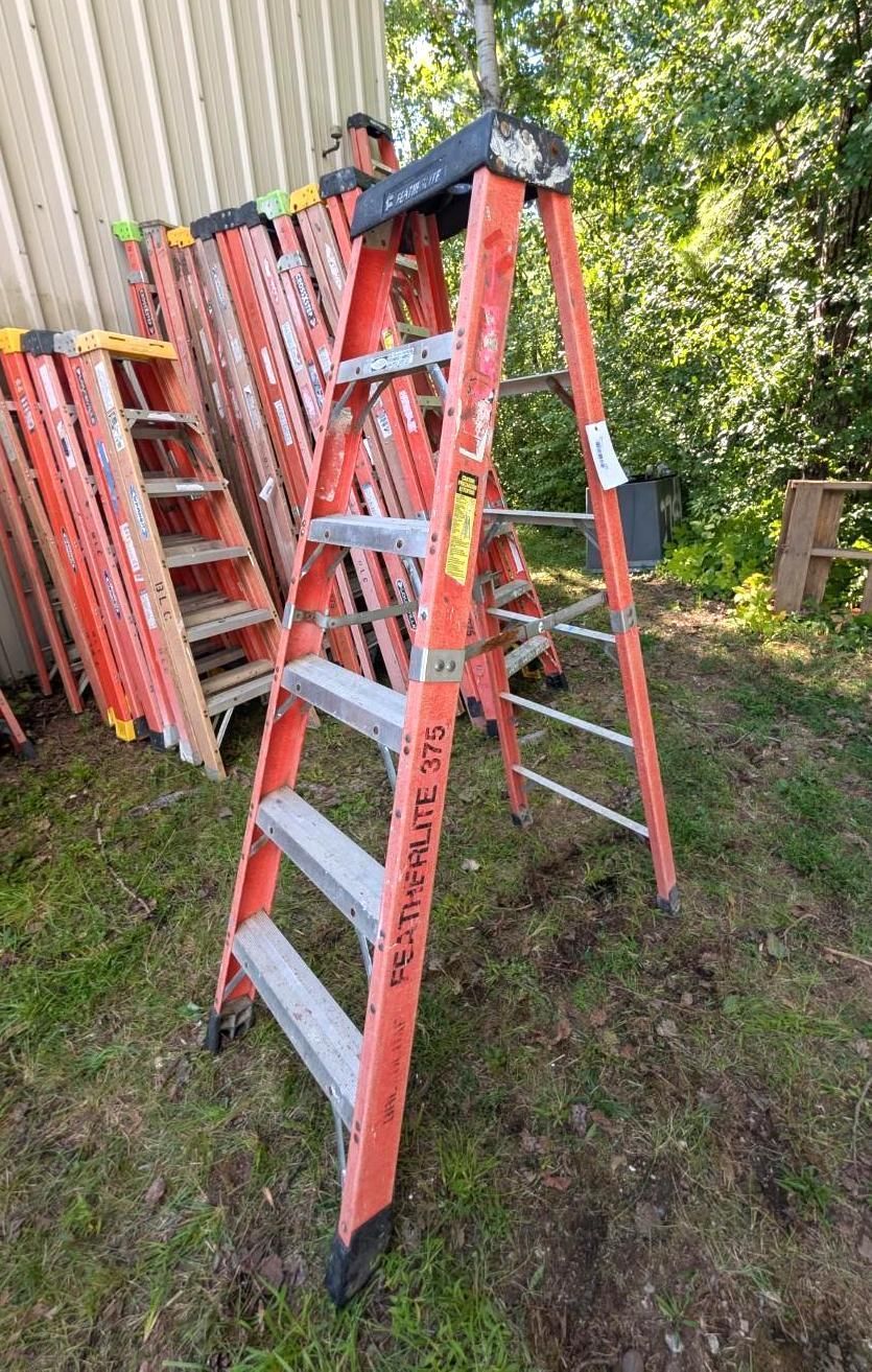 Red step ladder leaning in a grassy yard beside stacked ladders and a wooden fence