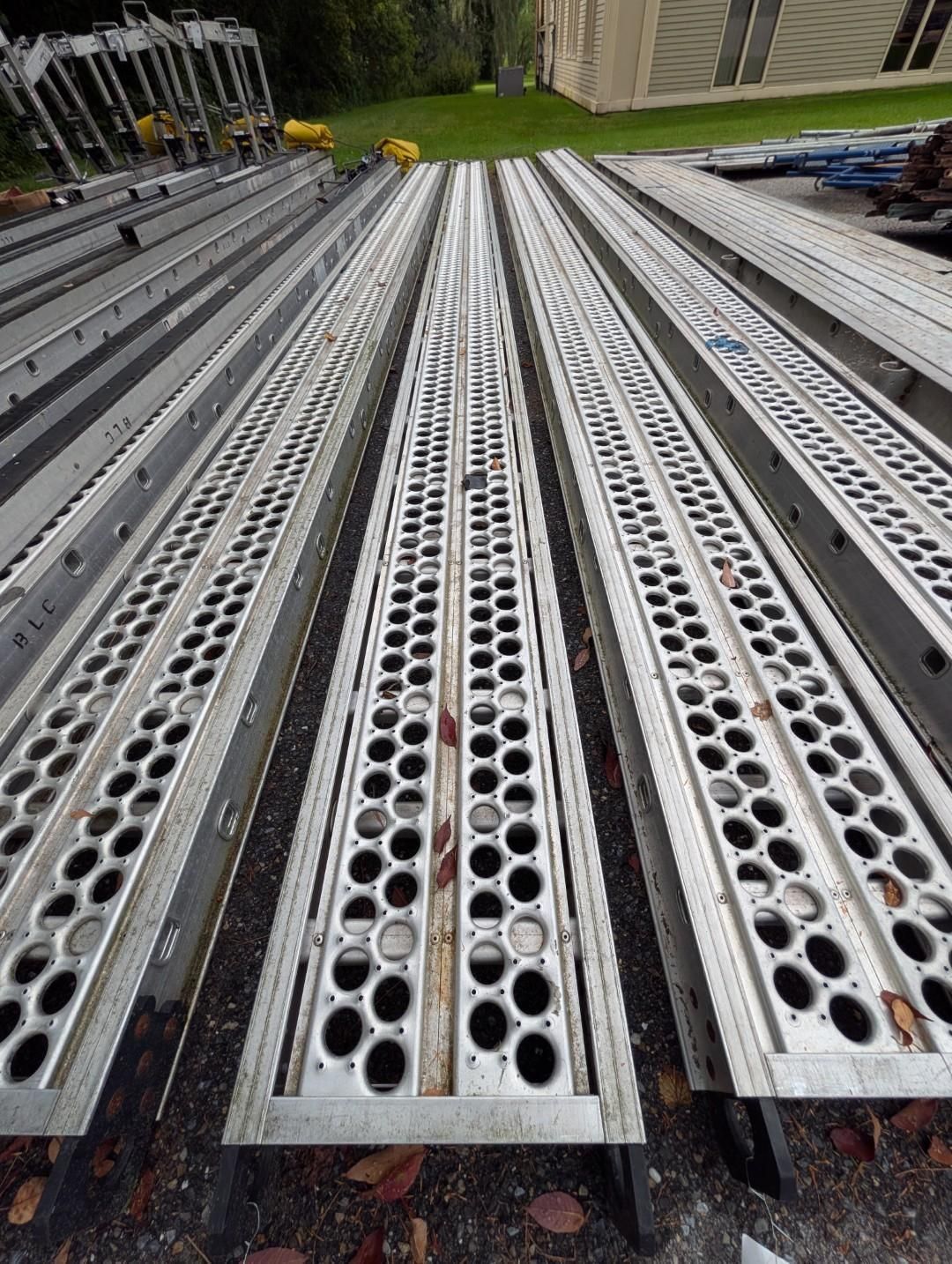 Rows of silver perforated metal scaffolding planks laid out outdoors on gravel