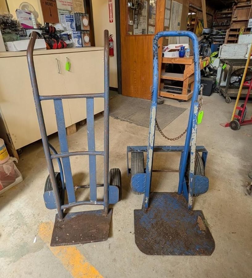 Two blue hand trucks on a workshop floor, one narrow and one wide, beside shelves and tools.