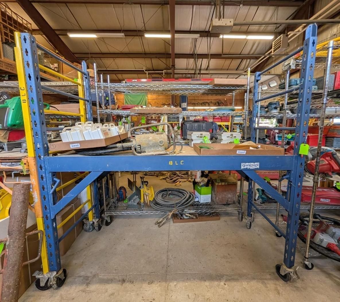 Warehouse workbench with blue metal rack and shelves of boxes, tools, and equipment under a high ceiling.