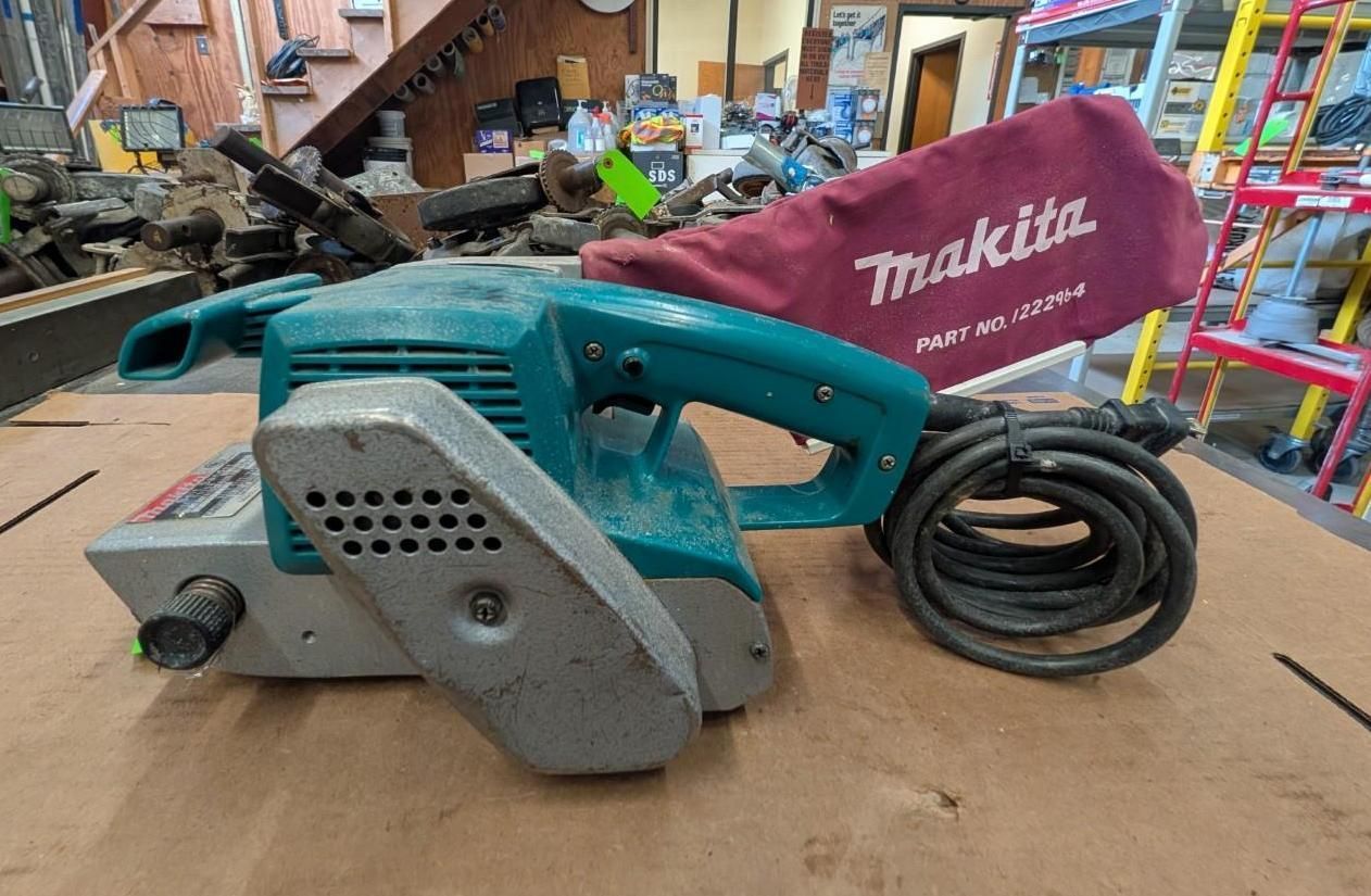 Makita electric belt sander with dust bag and cord on a workbench in a workshop