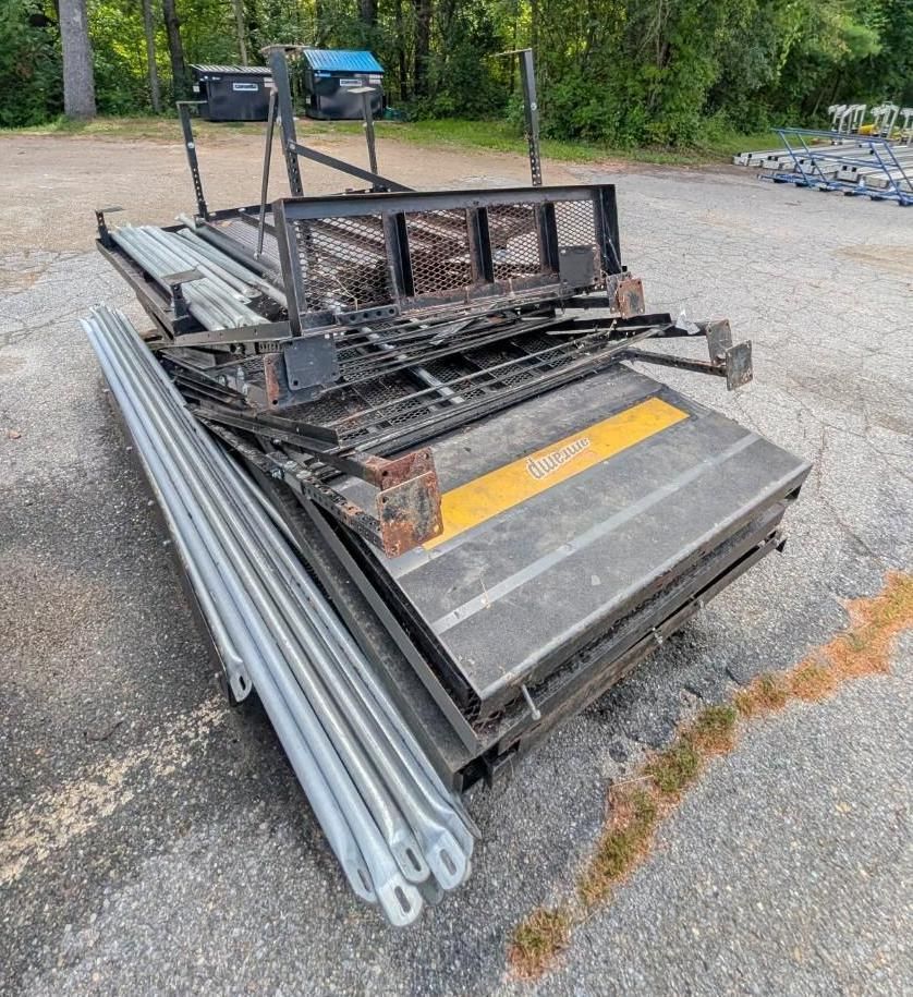 Stacked metal trailer parts and silver pipes on gravel in an outdoor yard.