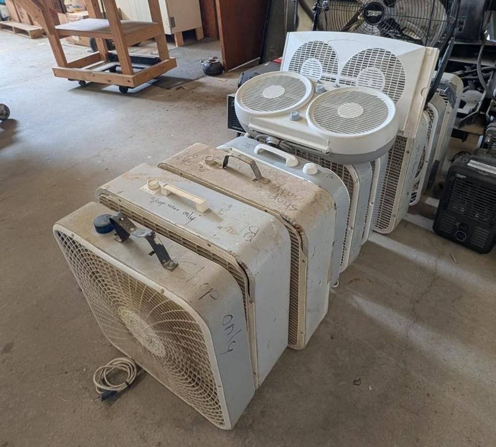 Stacked white box fans and small portable fans stored on a garage floor.