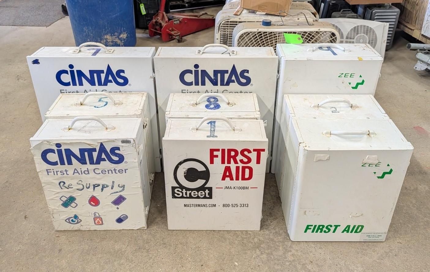 Six white and blue first aid coolers and boxes stacked on a concrete floor.